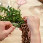 先以鐵絲穿過藤圈並收好，抓一把雪松，繞過後拉緊，再取一把雪松，在第一把根部接上，以此將雪松綁完。